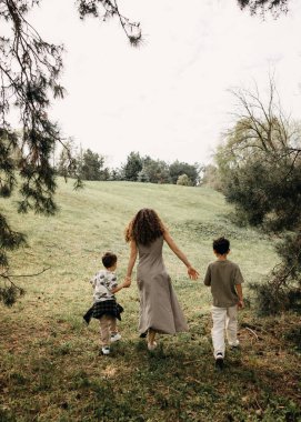 Anne ve iki oğlu güneşli bir orman çayırında yokuş yukarı yürüyorlar..