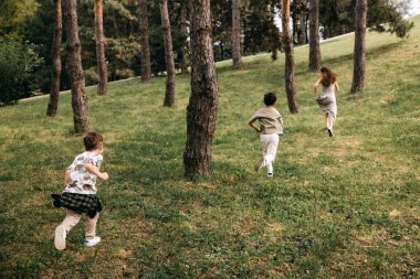 Anne ve iki çocuk parktaki uzun ağaçların arasında neşeyle koşuyorlar.