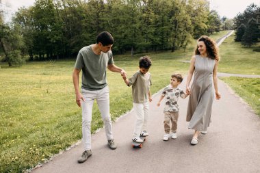 Aile çocuğu parkta kaykay sürmeyi öğretiyor, el ele tutuşuyorlar..