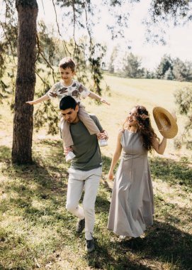 Gülümseyen çocuk babasının omuzlarında kollarını açmış güneşli yeşil parkta ailesiyle birlikte yürüyordu..