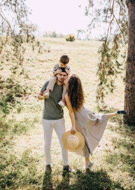 Anne bacağını kaldırırken baba, oğul omuzlarda, neşeli aile zamanı yeşil parkta.
