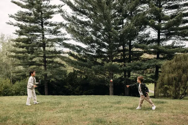Uzun çam ağaçlarının önünde çimde raket oynayan iki çocuk..