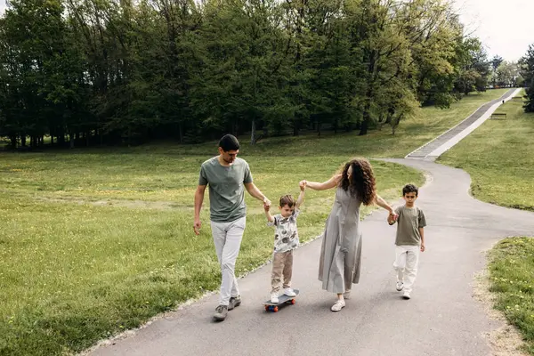 Aile parkta yürüyor, güneşli bir günde çocuğun kaykay sürmesine yardım ediyor..