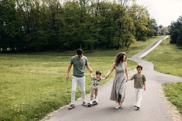 Aile parkta yürüyor, güneşli bir günde çocuğun kaykay sürmesine yardım ediyor..