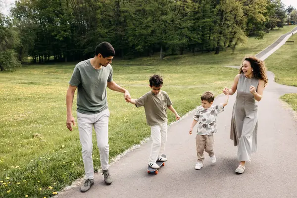 Mutlu bir aile parkta yürüyor, çocuklar birlikte kaykay öğreniyor..