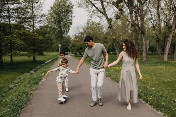 Aile çocuğu parkta kaykay sürmeyi öğretiyor, el ele tutuşuyorlar..