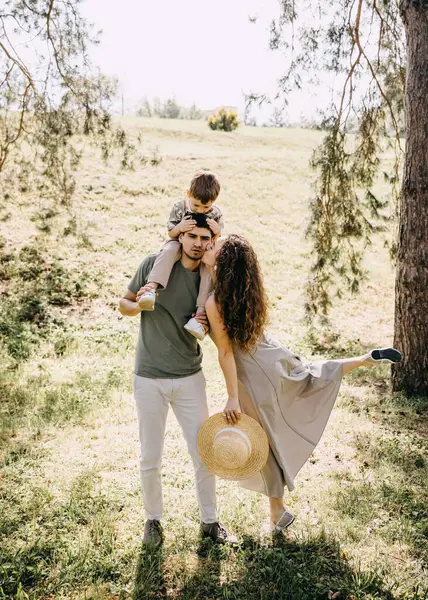 Anne bacağını kaldırırken baba, oğul omuzlarda, neşeli aile zamanı yeşil parkta.