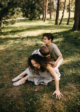 Annesi neşeyle gülerken iki küçük oğlu güneşli bir orman parkında neşe içinde üstüne çullanıyor..