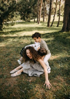Annesi neşeyle gülerken iki küçük oğlu güneşli bir orman parkında neşe içinde üstüne çullanıyor..