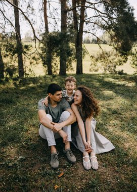 Küçük bir çocuk arkalarında neşeyle oynarken mutlu bir aile ormanda çimlerin üzerinde oturup birbirlerine gülümsüyor..