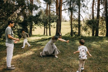 Aile ormanda göz bağı oyunu oynuyor, anne uzanıyor, çocuklar ve babalar güneşli bir günde gülüyor ve etkileşime geçiyor..