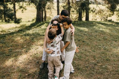 Çocuklar, güneşli bir günde parkta gülümseyen bir anneden göz bağını çıkarmaya yardım ediyor..