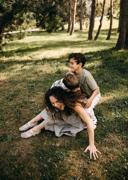 Annesi neşeyle gülerken iki küçük oğlu güneşli bir orman parkında neşe içinde üstüne çullanıyor..