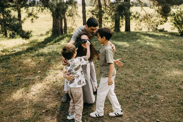 Çocuklar, güneşli bir günde parkta gülümseyen bir anneden göz bağını çıkarmaya yardım ediyor..