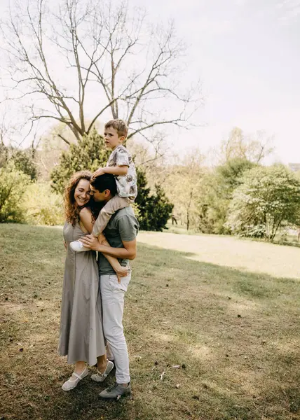 Dışarıda neşeli bir aile anı. Anne ve baba kucaklaşır. Oğul, babasının omuzlarında oturur. Güneşli bir parkta gülümserler..