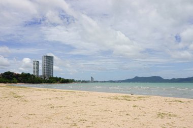 Beyaz bulutlarla dolu bir gökyüzünün altında durgun, gri-mavi bir denize açılan kumlu bir sahil..