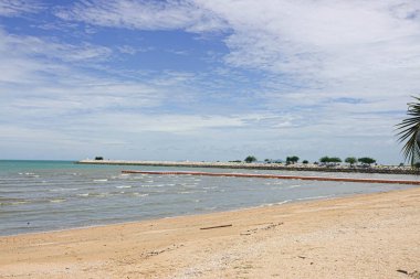 Beyaz bulutlarla dolu bir gökyüzünün altında durgun, gri-mavi bir denize açılan kumlu bir sahil..