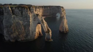 Güneş batarken, Etretat 'ın nefes kesen uçurumları çarpıcı siluetlerini canlı gökyüzüne karşı sergiliyor. Dalgalar eşsiz kaya oluşumlarına nazikçe yaklaşarak sakin bir sahil atmosferi oluşturur..