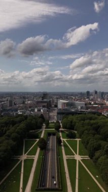 Parc du Cinquantenaire 'de sakin bir yolculuk sergileniyor, yemyeşil ve uzak şehrin ufuk çizgisi yumuşak bulutların altında, barışı ve yansımayı davet ediyor..