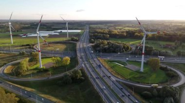 Güneş batarken, rüzgâr türbinleri ufukta dik duruyor, hareketli bir otoyolun üzerine gölgeler saçıyor. Doğa ve insan aktivitelerinin zıtlığı çarpıcı bir görsel uyum yaratır..