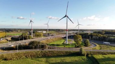Yükselen rüzgar türbinlerinin çarpıcı görüntüsü hareketli bir otobana karşı birleşerek temiz mavi gökyüzü altında doğanın ve modern altyapının uyumunu yakalıyor..