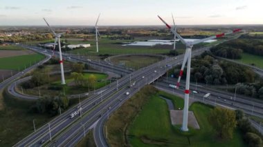 Görkemli rüzgar türbinleri işlek bir otoyol üzerinde doğayla teknolojiyi harmanlayarak yükseliyor. Araçlar, manzaranın önünden geçerken enerji ve hareketin ahenkli bir dengesini canlı bir şekilde gösteriyor..