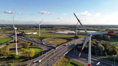 Doğanın ve teknolojinin, dağınık bulutlarla dolu canlı bir gökyüzü altında ahenkli bir şekilde bir arada var olduğu yoğun bir otoyolun yanında duran rüzgar türbinlerinin yükseldiği güzel bir manzara sergileniyor..