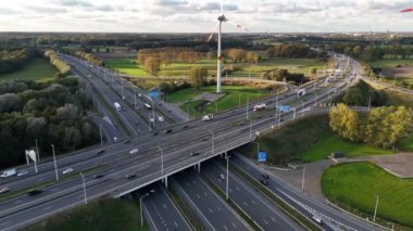 Yeşillik ve açık gökyüzü ile çevrili hareketli bir otoyolun üzerinde görkemli bir rüzgar türbini kuleleri. Otomobiller yol boyunca ilerler, doğanın uyumunu ve modern ulaşımı vurgularlar..