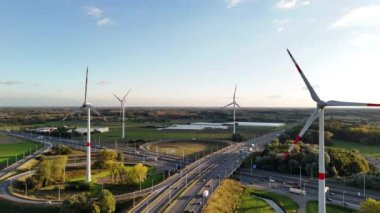 Dev rüzgâr türbinleri zarif bir şekilde dönerken araçlar otoyol boyunca ilerleyip yenilenebilir enerji ve hava kararırken manzarada seyahat etmenin uyumunu sergiliyor..