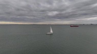 Sakin bir yat, kargo gemileriyle çevrili sakin sularda zarif bir şekilde gezinir. Kargo gemileri, karamsar bir gökyüzünün altında. Gün geceye dönüşürken, huzurlu bir deniz atmosferi yaratır..