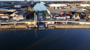 Büyük bir mavna, modern depo ve köprülerle çevrili sakin bir kanal boyunca sorunsuzca manevra yapıyor. Sakin su, berrak gökyüzünü yansıtır ve su yolunda sakin bir atmosfer yaratır..