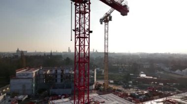 İşçiler şehrin ufuk çizgisi arasında koordinasyon sağlarken vinçler hareketli bir inşaat alanının üzerinde kule gibi çalışıyor. Gün ağarıyor, şehir gelişiminin gelişen manzarasına altın rengi yansıtıyor..