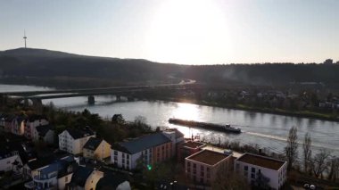 Güneş Koblenz üzerinde batarken Ren Nehri turuncu ve altın renklerini yansıtır. Şehir cazibesi ve uzak tepeler tarafından çerçevelenmiş, sakin bir atmosfer yaratan bir mavna..