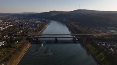 Alacakaranlık Koblenz üzerine inerken, Ren nehrinin sakin sularında yüzen bir mavnayı seyredin, manzaranın güzelliği ve uzak tepeler tarafından çerçevelenmiş..