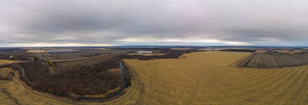 Drone panorama of autumn colored farm fields after harvest with a small river under a grey sky