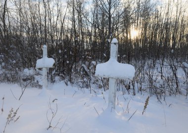 İki eski ahşap mezar taşı, arka planda ağaçlar ve ufukta güneş olan kalın bir kar tabakasıyla kaplı..