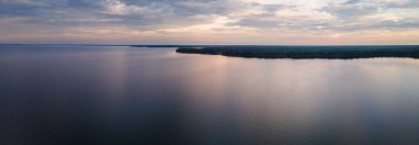 Gün doğumunda gökyüzü mavi, pembe ve gri bulutlarla dolu büyük göl manzarası. Suyun küçük dalgaları vardır ve gökyüzünden gelen renkleri yansıtır..