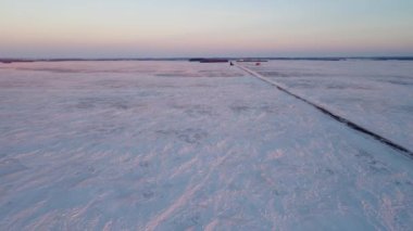 Gün batımına yakın, karla kaplı geniş bir bozkırın üzerindeki havadan. Düz bir yol tarlaları geçer. Kar ve gökyüzünün pembe bir tonu var..
