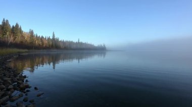 Sabahın erken saatlerinde, sağ tarafında sis olan karışık kuzey ağaçlarının bulunduğu sakin bir göl manzarası. Çok küçük dalgalar açık mavi gökyüzünü ve ağaçları yansıtıyor..  