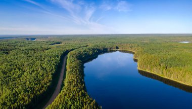 Büyük yeşil bir ormanın parlak bir yaz havası ve ince beyaz bulutlu mavi bir gökyüzünün altında sakin bir göl. Bir çakıllı yol ağaçların arasından geçer ve göl kıyısını takip eder. Sakin su, gökyüzünün mavi rengini yansıtır..