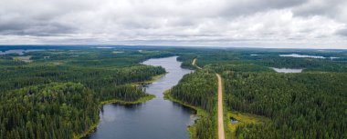 Birçok gölü ve uzun çakıl yolu olan uçsuz bucaksız bir ormanın havadan manzarası. Ormanda çoğunlukla çam ağaçları ve çam ağaçları var. Yol uzun bir gölün kıyısını takip ediyor. Gökyüzü gri bulutlarla dolu..
