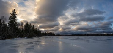 Donmuş bir gölde karamsar bir kış sahnesi. Kıyı şeridi ladin ve yaprak döken ağaçların karışımıdır. Parlak buzun üzerinde kar yok ve gökyüzü çok renkli bulutlarla dolu..  