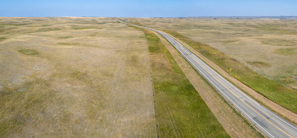 Вид с воздуха, смотрящий вдоль двухполосной асфальтированной сельской автомагистрали, которая пересекает и открывает безлесные прерии и исчезает вдали. Небо светло-голубое, облаков нет.  