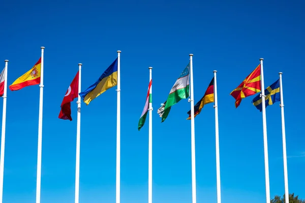 Flags of Different Countries Hang On Flagpoles Against Blue Sky. Flag of Spain, Ukraine, Germany, Sweden, Turkey, Uzbekistan and Macedonia. The concept of support and unity of countries. High quality