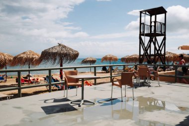 Moraitika, Greece - 09 27 2022: People Are Sunbathing On Sunbeds Near Water Of Ionian Sea, In Foreground - Terrace Of Beach Cafe Wet After Rain. High quality photo clipart