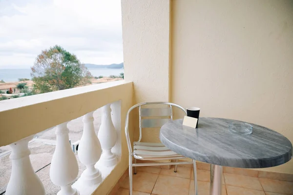 In Frame There Is Greek Hotel Balcony With Metal Chair And Stone Table, Glass Ashtray, Bank Card And Paper Cup Of Coffee On Table. Concept Of Payment For Vacation. High quality photo