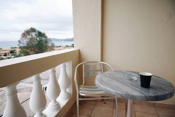 In Frame There Is Greek Hotel Balcony With Metal Chair And Stone Table, Glass Ashtray And Paper Cup Of Coffee On Table. High quality photo