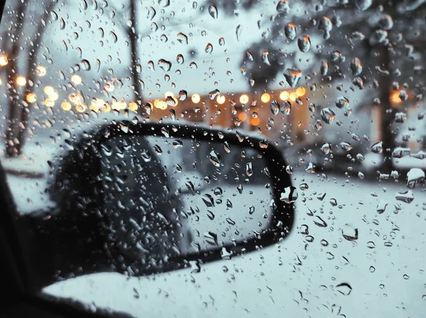 Close-Up Of Water Droplets On Side Window Of Car During Snowfall In Winter, Against Background Of Rearview Mirror And Bokeh Of Street Lights. High quality photo