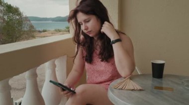 Young Woman In Bright Dress Is Sitting On Chair On Hotel Balcony, Holding Phone, touches her hair And and enjoying the moment. On Table Fan, Cup Of Tea And Gold Card. . High quality 4k footage