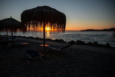 Tatil köyünde şafak sökerken, güneş ışınları plaj şemsiyesini palmiye yapraklarından kırıyor. Plaj sandalyeleri, şemsiyeler ve iskele siluetleri. Yüksek kalite fotoğraf